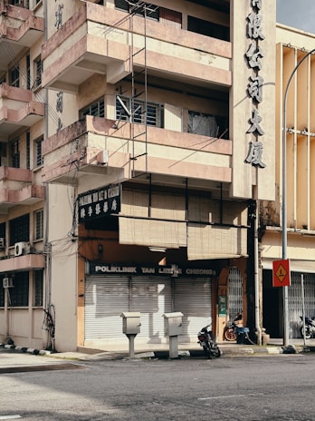 A multi-story building with a sun-worn façade and balconies rising above a closed clinic named 'Poliklinik Tan Lee & Cheong'. The building's exterior displays large Chinese characters, and wires run along the walls. Below, two motorcycles are parked beside the entrance, and a few utility boxes are positioned near the street corner. A traffic sign stands nearby, indicating a sharp turn. The environment appears urban with minimal activity.