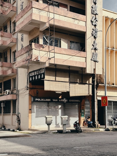 A multi-story building with a sun-worn façade and balconies rising above a closed clinic named 'Poliklinik Tan Lee & Cheong'. The building's exterior displays large Chinese characters, and wires run along the walls. Below, two motorcycles are parked beside the entrance, and a few utility boxes are positioned near the street corner. A traffic sign stands nearby, indicating a sharp turn. The environment appears urban with minimal activity.