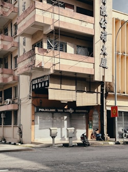 A multi-story building with a sun-worn fa&ccedil;ade and balconies rising above a closed clinic named 'Poliklinik Tan Lee & Cheong'. The building's exterior displays large Chinese characters, and wires run along the walls. Below, two motorcycles are parked beside the entrance, and a few utility boxes are positioned near the street corner. A traffic sign stands nearby, indicating a sharp turn. The environment appears urban with minimal activity.