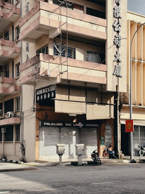 A multi-story building with a sun-worn façade and balconies rising above a closed clinic named 'Poliklinik Tan Lee & Cheong'. The building's exterior displays large Chinese characters, and wires run along the walls. Below, two motorcycles are parked beside the entrance, and a few utility boxes are positioned near the street corner. A traffic sign stands nearby, indicating a sharp turn. The environment appears urban with minimal activity.