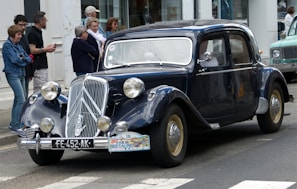 A vintage black car parked on a street, surrounded by a group of people standing on the sidewalk. The car features classic design elements such as round headlights and a distinctive chrome grille. A number plate and a tour sticker are visible on the front. The background includes a storefront and another vehicle partially visible.