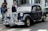 A vintage black car parked on a street, surrounded by a group of people standing on the sidewalk. The car features classic design elements such as round headlights and a distinctive chrome grille. A number plate and a tour sticker are visible on the front. The background includes a storefront and another vehicle partially visible.