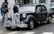 A vintage black car parked on a street, surrounded by a group of people standing on the sidewalk. The car features classic design elements such as round headlights and a distinctive chrome grille. A number plate and a tour sticker are visible on the front. The background includes a storefront and another vehicle partially visible.