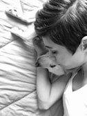 A person with short hair is lying down, closely embracing a dog, creating an intimate and comforting scene. They both appear relaxed on a soft, quilted surface.
