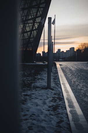 A modern architectural structure with glass facades stands prominently. Tall flag poles are aligned along a pathway, with a light dusting of snow on the ground. The sun is setting in the background, casting an orange glow over an urban skyline of high-rise buildings.