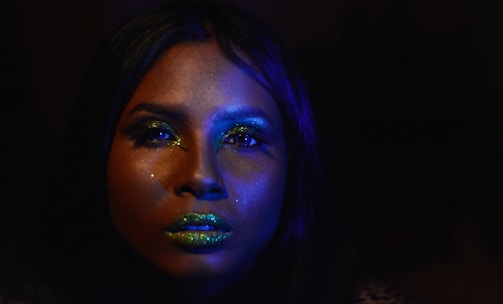 A close-up of a person with shimmering makeup, featuring glittery green lips and eyeshadow. The lighting casts an ethereal blue glow across the face, accentuating the shine.