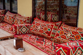 A wooden table with a tissue box and an ashtray is placed in front of a seating area adorned with vibrant, patterned cushions and blankets. The cushions have intricate geometric designs mostly in red, green, and white colors. The seating area is positioned next to a glass window with reflections of the cushions and blankets visible.