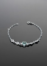 A delicate silver bracelet with a central heart-shaped gemstone set in the middle. The bracelet features several small round stones arranged symmetrically on either side of the heart. It is laid out on a smooth, dark surface that enhances the shine and elegance of the jewelry.