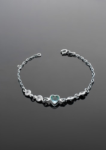 A delicate silver bracelet with a central heart-shaped gemstone set in the middle. The bracelet features several small round stones arranged symmetrically on either side of the heart. It is laid out on a smooth, dark surface that enhances the shine and elegance of the jewelry.