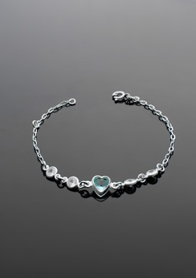 A delicate silver bracelet with a central heart-shaped gemstone set in the middle. The bracelet features several small round stones arranged symmetrically on either side of the heart. It is laid out on a smooth, dark surface that enhances the shine and elegance of the jewelry.