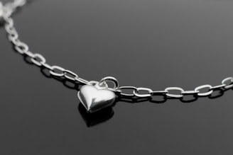 A close-up of a delicate silver necklace with a small heart pendant on a soft white fabric background.