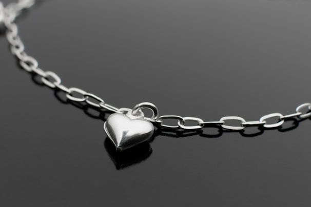 A close-up of a delicate silver necklace with a small heart pendant on a soft white fabric background.