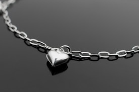 A delicate silver chain necklace with a polished heart-shaped pendant against a dark background. The chain features interlocking oval links, and the pendant is centrally positioned, reflecting light and adding a touch of elegance.