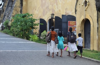 Four people, including a woman and three children, walk along a paved path towards a structure with a yellowish wall and dark doors. The individuals carry backpacks and one of the children holds a pink item. Lush greenery lines the path, and a red and yellow entrance sign is visible near the building.