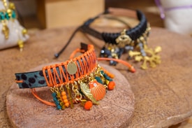 Intricately designed bracelets featuring vibrant colors and dangling ornaments rest on a textured wooden surface. One bracelet is woven with orange thread, showcasing gold charms and colorful beads, while the other has a black base with similar gold details.