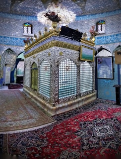 An ornate interior featuring an intricately designed tomb or shrine surrounded by an elaborate silver lattice structure with floral arrangements on top. The walls are adorned with detailed blue and gold calligraphy, and the space is enriched with colorful stained glass windows. Persian carpets with complex patterns cover the floor.