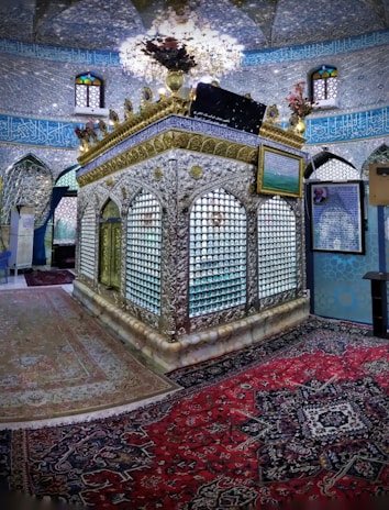 An ornate interior featuring an intricately designed tomb or shrine surrounded by an elaborate silver lattice structure with floral arrangements on top. The walls are adorned with detailed blue and gold calligraphy, and the space is enriched with colorful stained glass windows. Persian carpets with complex patterns cover the floor.