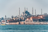 A scenic view of a large mosque with multiple minarets situated on a hill, surrounded by a mix of residential and historical buildings. A prominent lighthouse and a yellow ferry can be seen in the foreground, against the backdrop of a calm blue sea.