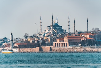 A scenic view of a large mosque with multiple minarets situated on a hill, surrounded by a mix of residential and historical buildings. A prominent lighthouse and a yellow ferry can be seen in the foreground, against the backdrop of a calm blue sea.