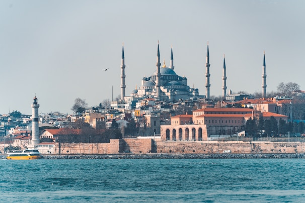 A scenic view of a large mosque with multiple minarets situated on a hill, surrounded by a mix of residential and historical buildings. A prominent lighthouse and a yellow ferry can be seen in the foreground, against the backdrop of a calm blue sea.