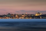 A panoramic view of Istanbul's skyline at dusk, featuring the illuminated silhouettes of historical landmarks against a soft pink and blue sky. The city lights reflect softly on the water in the foreground, adding a sense of calm and beauty.
