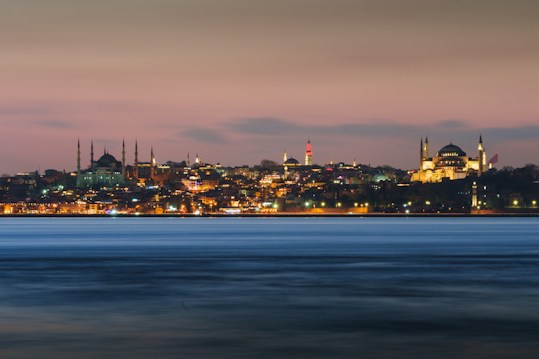 A panoramic view of Istanbul's skyline at dusk, featuring the illuminated silhouettes of historical landmarks against a soft pink and blue sky. The city lights reflect softly on the water in the foreground, adding a sense of calm and beauty.