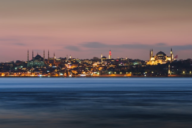 A panoramic view of Istanbul's skyline at dusk, featuring the illuminated silhouettes of historical landmarks against a soft pink and blue sky. The city lights reflect softly on the water in the foreground, adding a sense of calm and beauty.
