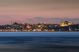 A panoramic view of Istanbul's skyline at dusk, featuring the illuminated silhouettes of historical landmarks against a soft pink and blue sky. The city lights reflect softly on the water in the foreground, adding a sense of calm and beauty.