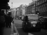 A busy urban street scene with a classic black taxi driving down the road. Pedestrians are walking on the sidewalk, and traffic signals are visible. The architecture of the surrounding buildings is historic and ornate, with multiple stories and intricate details.