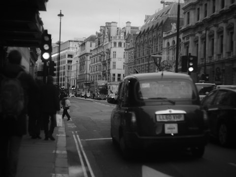 A busy urban street scene with a classic black taxi driving down the road. Pedestrians are walking on the sidewalk, and traffic signals are visible. The architecture of the surrounding buildings is historic and ornate, with multiple stories and intricate details.