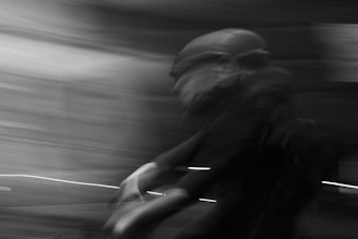 A motion blur effect captures a person on a bicycle, wearing a helmet and moving rapidly through a dimly lit environment. The background is a mix of swirling lines and shadows, suggesting speed and movement.