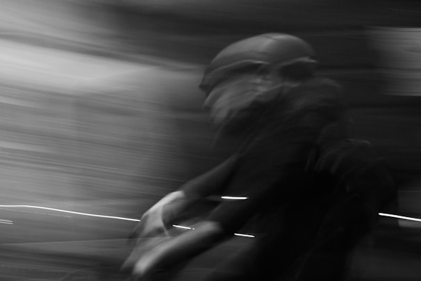 A motion blur effect captures a person on a bicycle, wearing a helmet and moving rapidly through a dimly lit environment. The background is a mix of swirling lines and shadows, suggesting speed and movement.