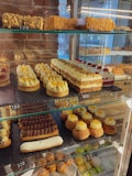 A bakery display case filled with a variety of desserts and pastries, including layered cakes, tarts topped with cream and fruit, chocolate éclairs, and a selection of macarons. The items are neatly arranged on multiple glass shelves, with some price tags visible.