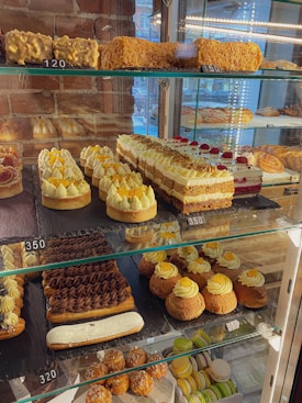 A bakery display case filled with a variety of desserts and pastries, including layered cakes, tarts topped with cream and fruit, chocolate éclairs, and a selection of macarons. The items are neatly arranged on multiple glass shelves, with some price tags visible.