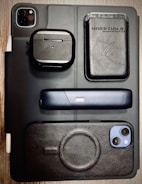 A flat lay of various electronic and personal items arranged on a surface. Items include a black iPad with dual camera lens, a pair of wireless earphones in a black case, a sleek black electronic device with a single button, a smartphone with a black case, and a black leather wallet with cards visible inside. The background is a wooden table, and the overall styling is modern and minimalist.