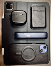 A flat lay of various electronic and personal items arranged on a surface. Items include a black iPad with dual camera lens, a pair of wireless earphones in a black case, a sleek black electronic device with a single button, a smartphone with a black case, and a black leather wallet with cards visible inside. The background is a wooden table, and the overall styling is modern and minimalist.