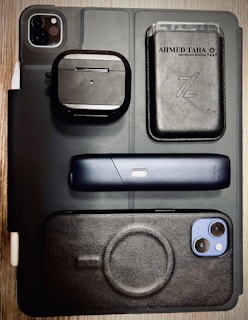 A flat lay of various electronic and personal items arranged on a surface. Items include a black iPad with dual camera lens, a pair of wireless earphones in a black case, a sleek black electronic device with a single button, a smartphone with a black case, and a black leather wallet with cards visible inside. The background is a wooden table, and the overall styling is modern and minimalist.