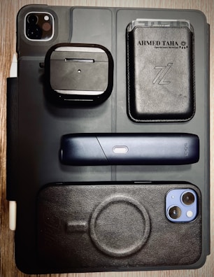 A flat lay of various electronic and personal items arranged on a surface. Items include a black iPad with dual camera lens, a pair of wireless earphones in a black case, a sleek black electronic device with a single button, a smartphone with a black case, and a black leather wallet with cards visible inside. The background is a wooden table, and the overall styling is modern and minimalist.