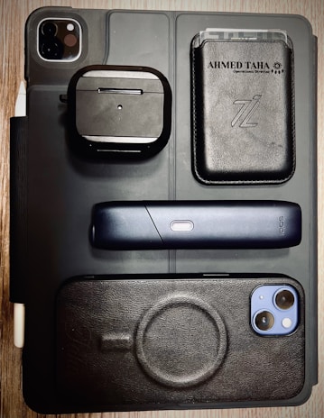 A flat lay of various electronic and personal items arranged on a surface. Items include a black iPad with dual camera lens, a pair of wireless earphones in a black case, a sleek black electronic device with a single button, a smartphone with a black case, and a black leather wallet with cards visible inside. The background is a wooden table, and the overall styling is modern and minimalist.