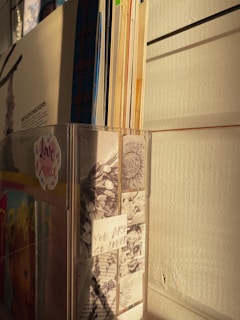 A collection of vinyl records is neatly organized in a vertical holder against a wooden wall. The front of the holder displays a collage of art, including illustrations and written notes, with a prominent message that reads 'You Are So Loved.' The setting is bathed in warm, golden light, suggesting either sunrise or sunset.
