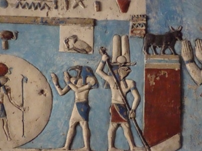 An ancient Egyptian relief featuring hieroglyphics and figures adorned in traditional garments and headpieces. The scene includes a figure with the head of a bird holding a staff, and a bull standing on a pedestal. The background includes blue and earthy tones, with detailed carvings depicting hands and other symbols.