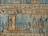 An ancient Egyptian relief featuring detailed hieroglyphics and figures on a background with a dominant blue tone. The central figure, possibly a deity, stands on a decorated boat adorned with smaller figures. To the right, two larger figures stand upright, also adorned in traditional Egyptian attire. The background is filled with star motifs.