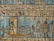 An ancient Egyptian relief featuring detailed hieroglyphics and figures on a background with a dominant blue tone. The central figure, possibly a deity, stands on a decorated boat adorned with smaller figures. To the right, two larger figures stand upright, also adorned in traditional Egyptian attire. The background is filled with star motifs.