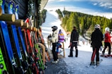 A group of people in ski outfits is gathered near a ski rental shop with various colorful skis lined up against the wooden exterior. In the background, a snow-covered ski slope stretches up a hill surrounded by dense trees under a bright blue sky, with additional skiers visible on the slope.