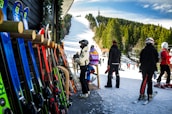 A group of people in ski outfits is gathered near a ski rental shop with various colorful skis lined up against the wooden exterior. In the background, a snow-covered ski slope stretches up a hill surrounded by dense trees under a bright blue sky, with additional skiers visible on the slope.