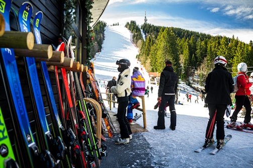 A group of people in ski outfits is gathered near a ski rental shop with various colorful skis lined up against the wooden exterior. In the background, a snow-covered ski slope stretches up a hill surrounded by dense trees under a bright blue sky, with additional skiers visible on the slope.