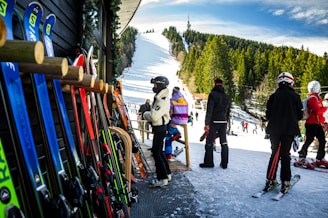 A group of people in ski outfits is gathered near a ski rental shop with various colorful skis lined up against the wooden exterior. In the background, a snow-covered ski slope stretches up a hill surrounded by dense trees under a bright blue sky, with additional skiers visible on the slope.