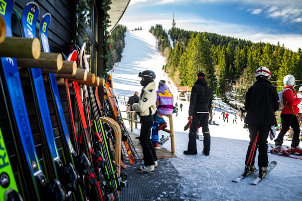 A group of people in ski outfits is gathered near a ski rental shop with various colorful skis lined up against the wooden exterior. In the background, a snow-covered ski slope stretches up a hill surrounded by dense trees under a bright blue sky, with additional skiers visible on the slope.