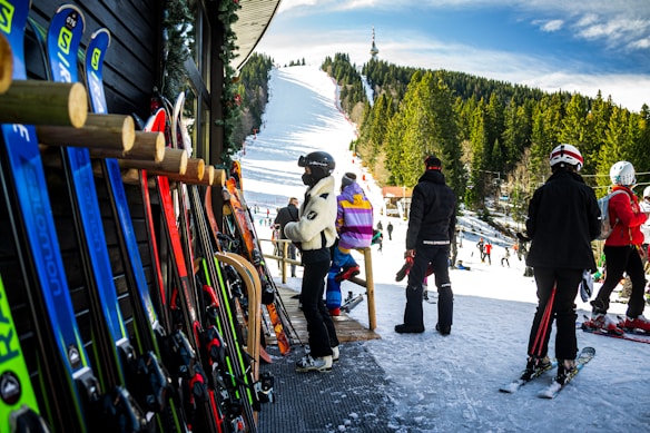 A group of people in ski outfits is gathered near a ski rental shop with various colorful skis lined up against the wooden exterior. In the background, a snow-covered ski slope stretches up a hill surrounded by dense trees under a bright blue sky, with additional skiers visible on the slope.