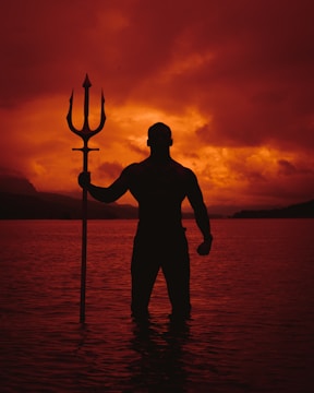 A silhouette of a muscular figure holding a trident stands partially submerged in dark water, with a dramatic, fiery sunset casting red and orange hues across the sky and reflecting on the water.